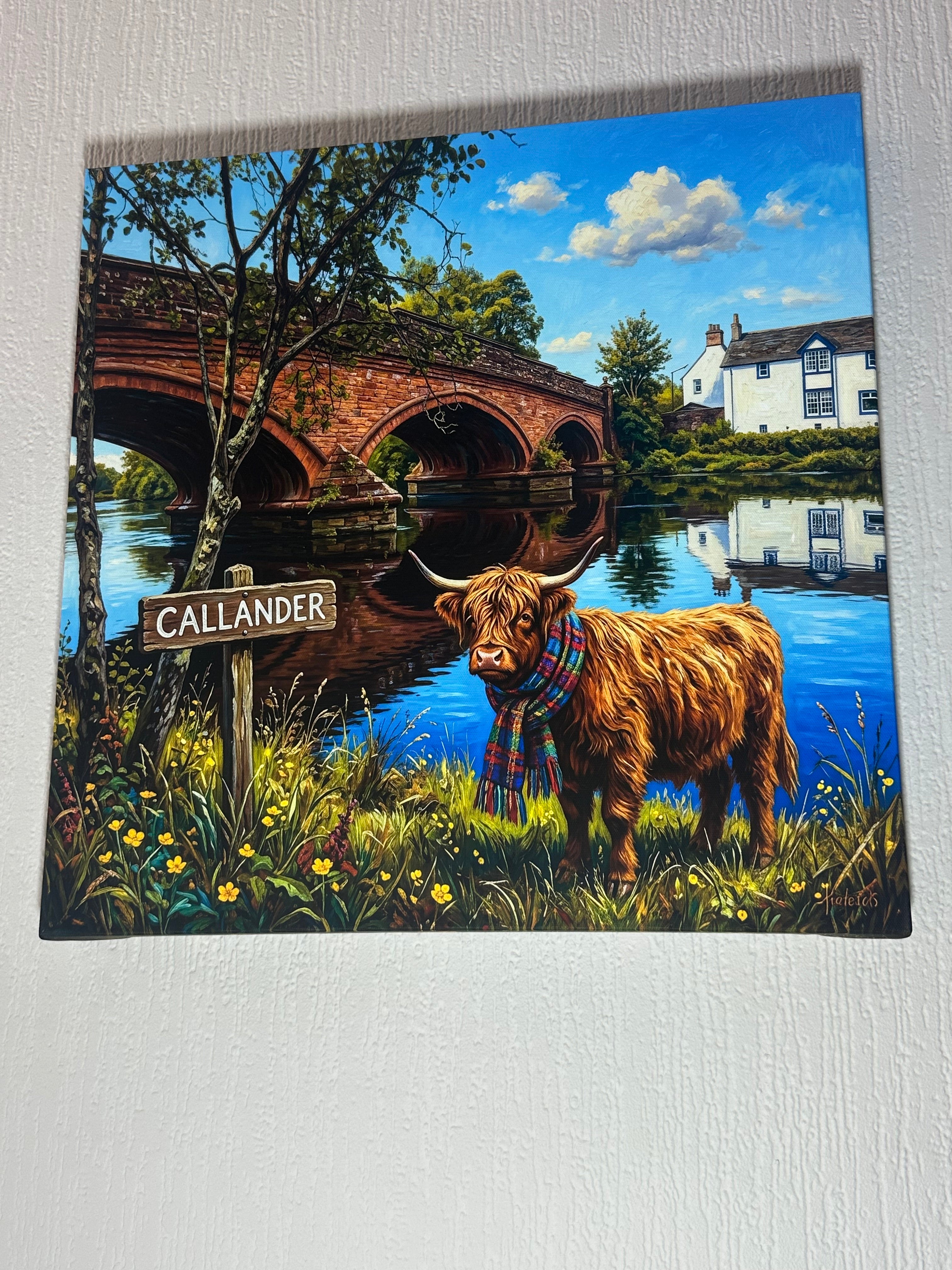 A square canvas print featuring a colorful, whimsical landscape of Scotland with a Highland cow in a plaid scarf, a brick bridge, a white house, and a 'CALLANDER' sign, displayed on a textured white wall.