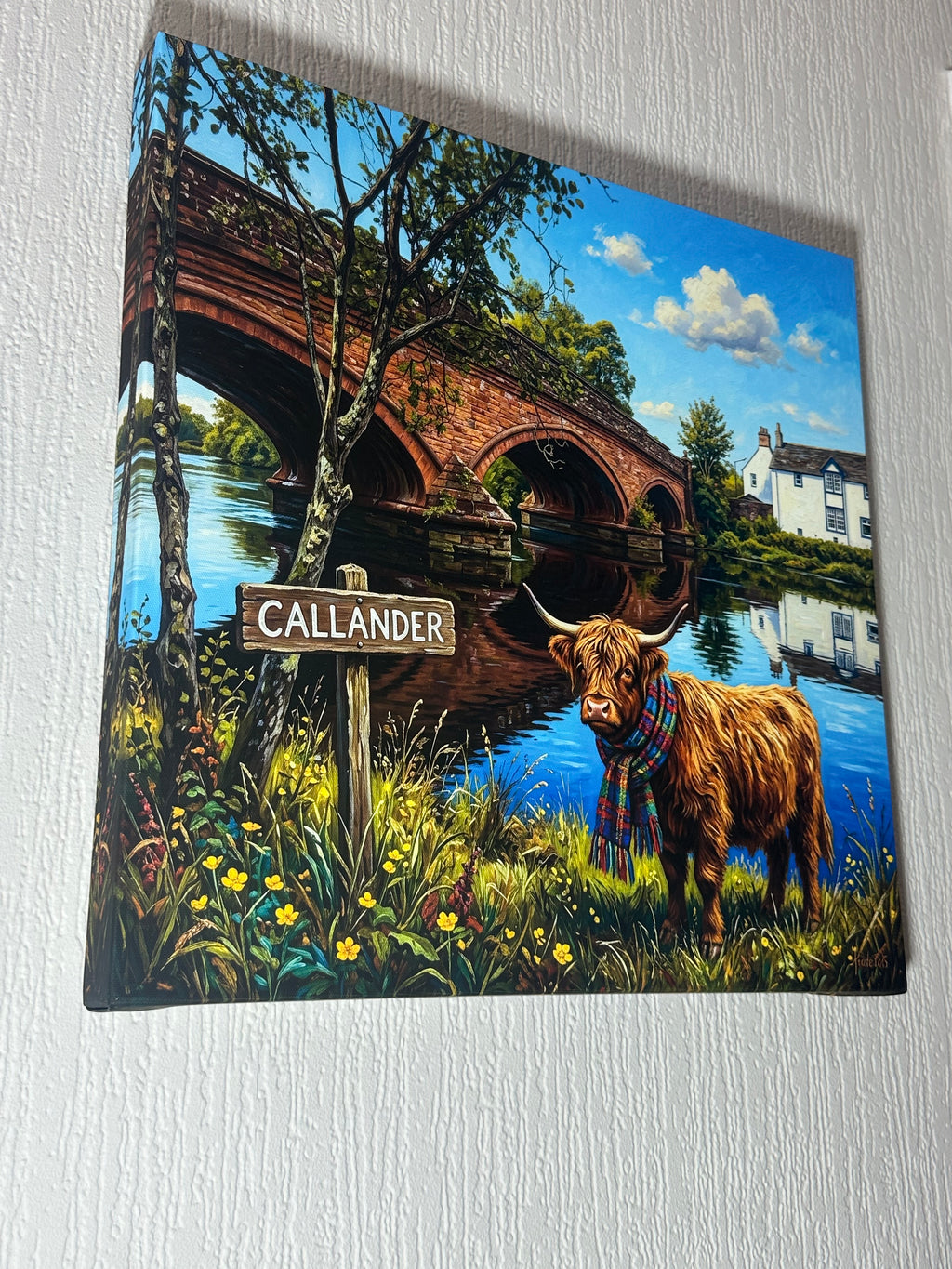 A square canvas print featuring a whimsical illustration of a Highland cow wearing a plaid kilt standing by a bridge with the sign 'CALLANDER', displayed on a textured white wall.