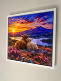 A framed wall art print showing two Highland cows sitting on a hillside with a colorful sunset over a lake and mountains, mounted on a light textured wall.