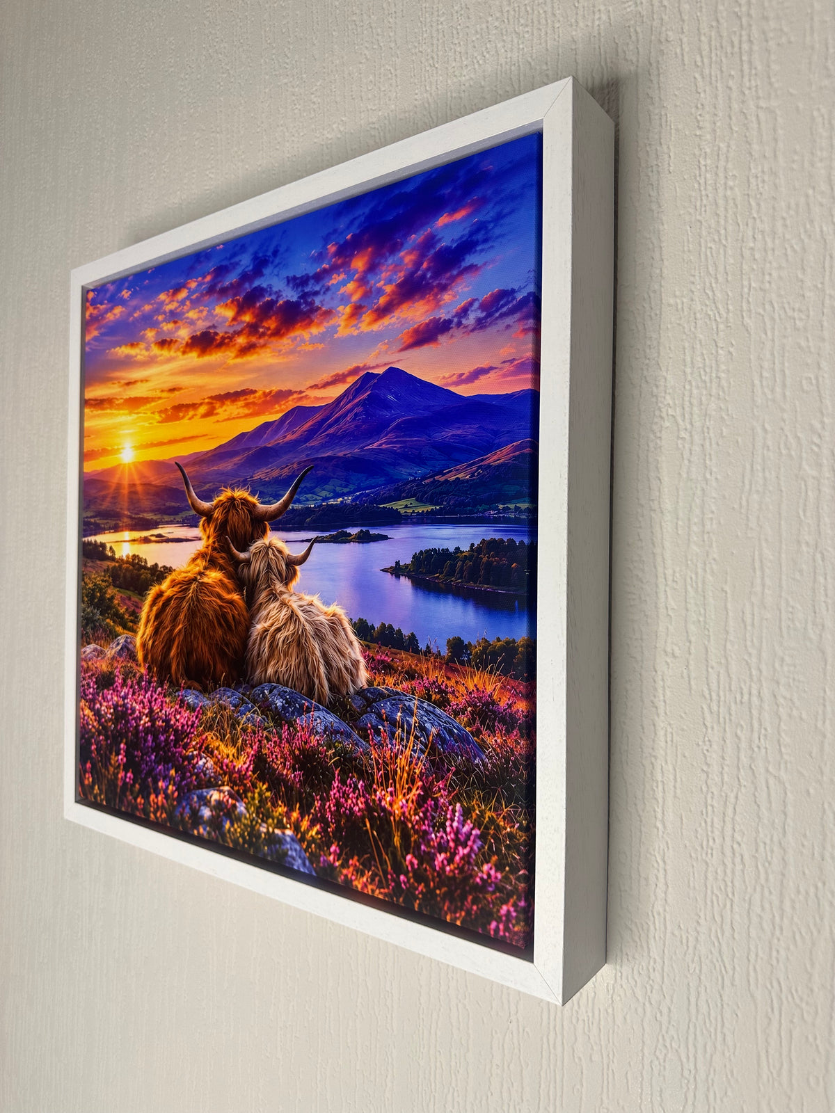 A framed wall art print showing two Highland cows sitting in a field of pink flowers, with a lake, mountain, and dramatic sunset sky in the background. The print is mounted in a white frame and displayed on a light textured wall.
