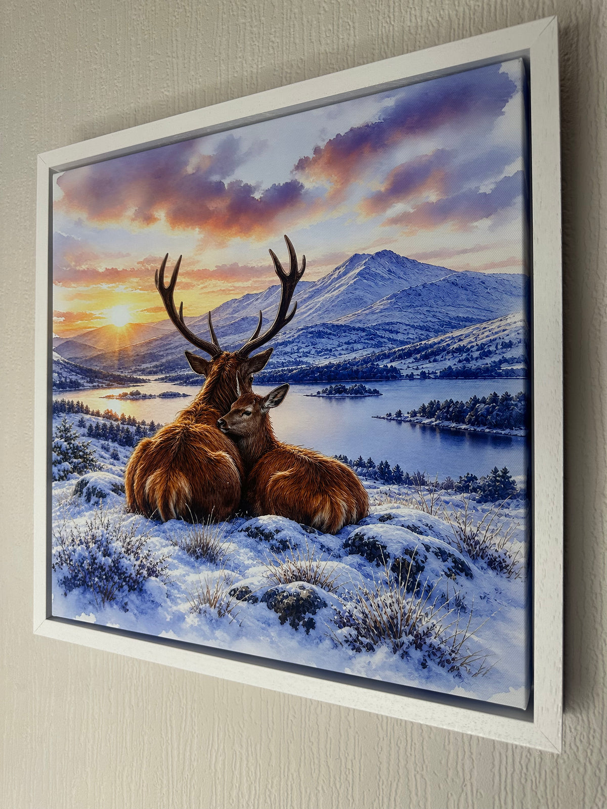 A framed wall art print showing two deer resting in a snowy landscape with mountains, a lake, and a dramatic sunset sky, mounted on a light beige textured wall.