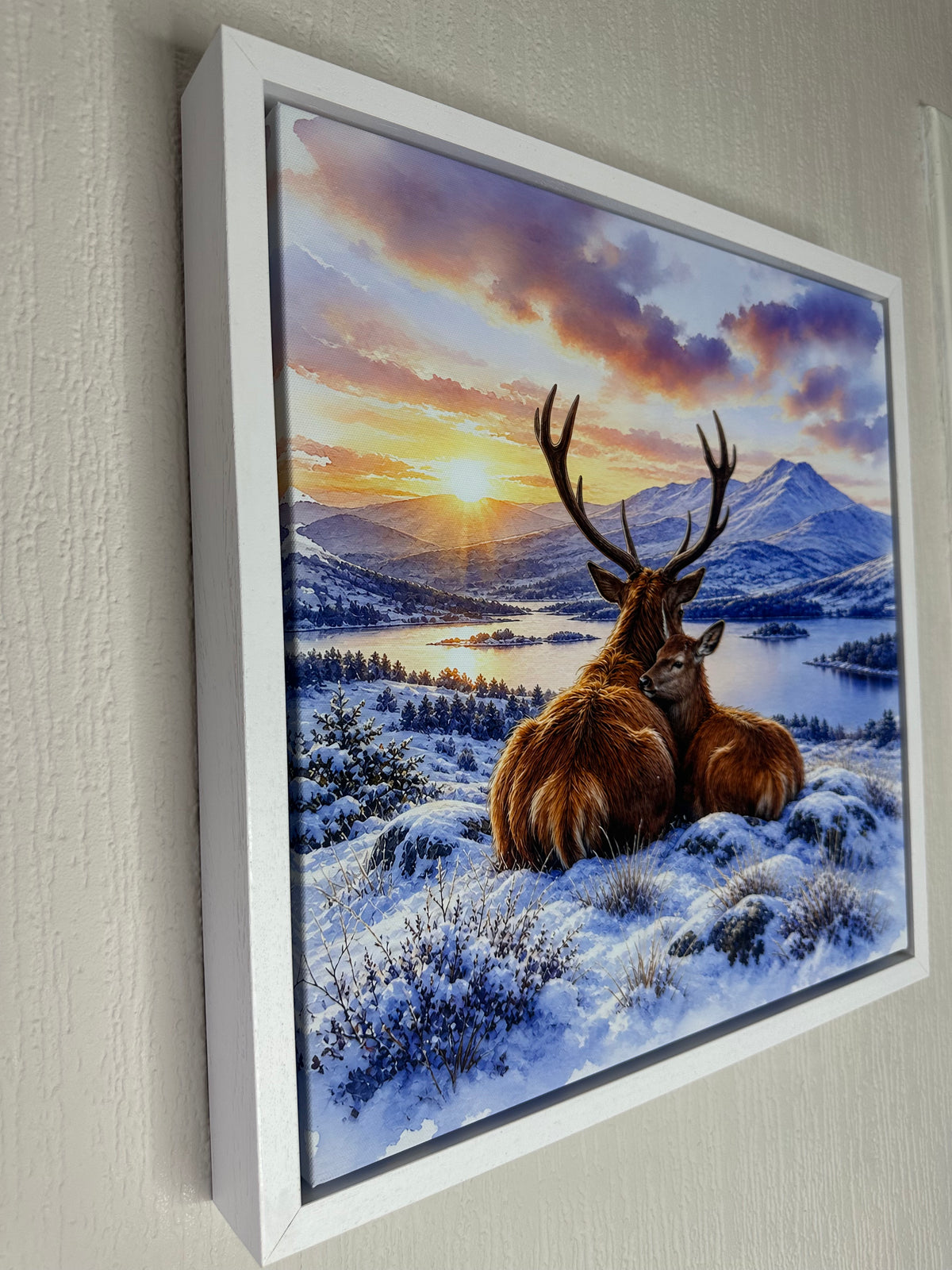 A framed wall art print showing a snowy winter landscape with a large brown deer and a smaller deer in the foreground, mountains, a lake, and a colorful sunset sky. The artwork is mounted on a light beige wall.