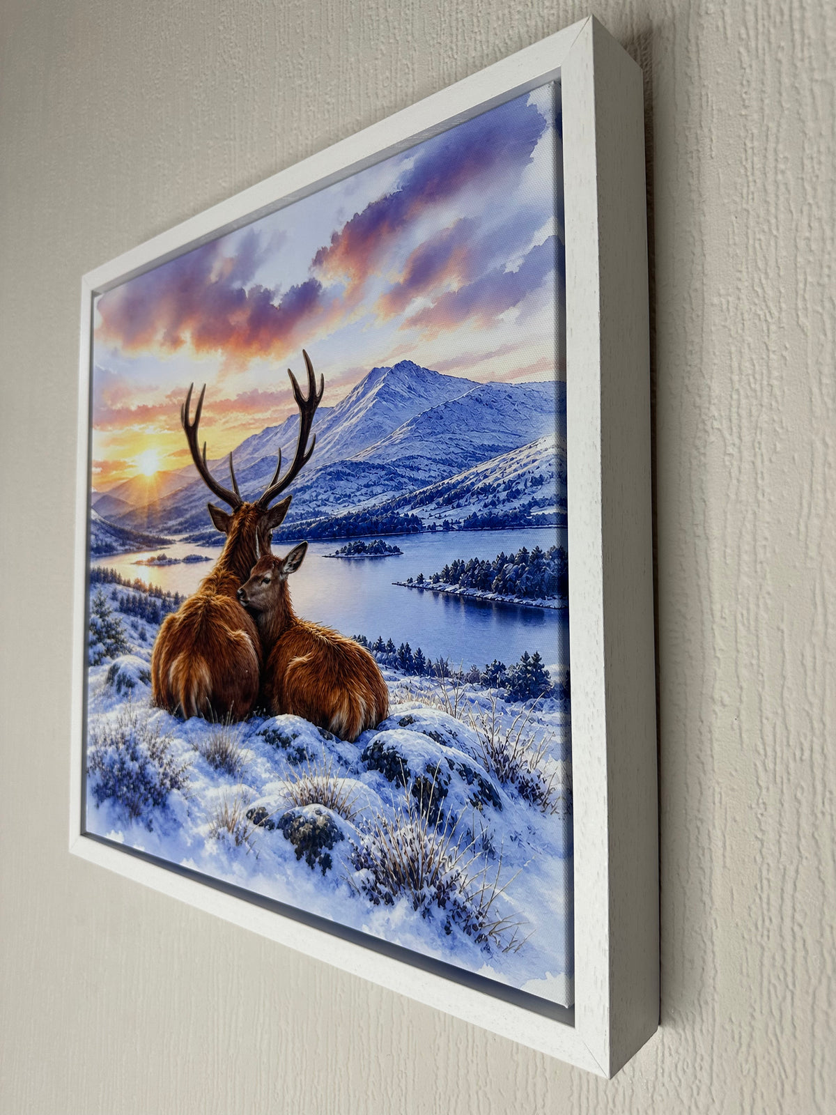 Framed wall art showing a snowy mountain landscape with two deer in the foreground, displayed against a light textured wall.