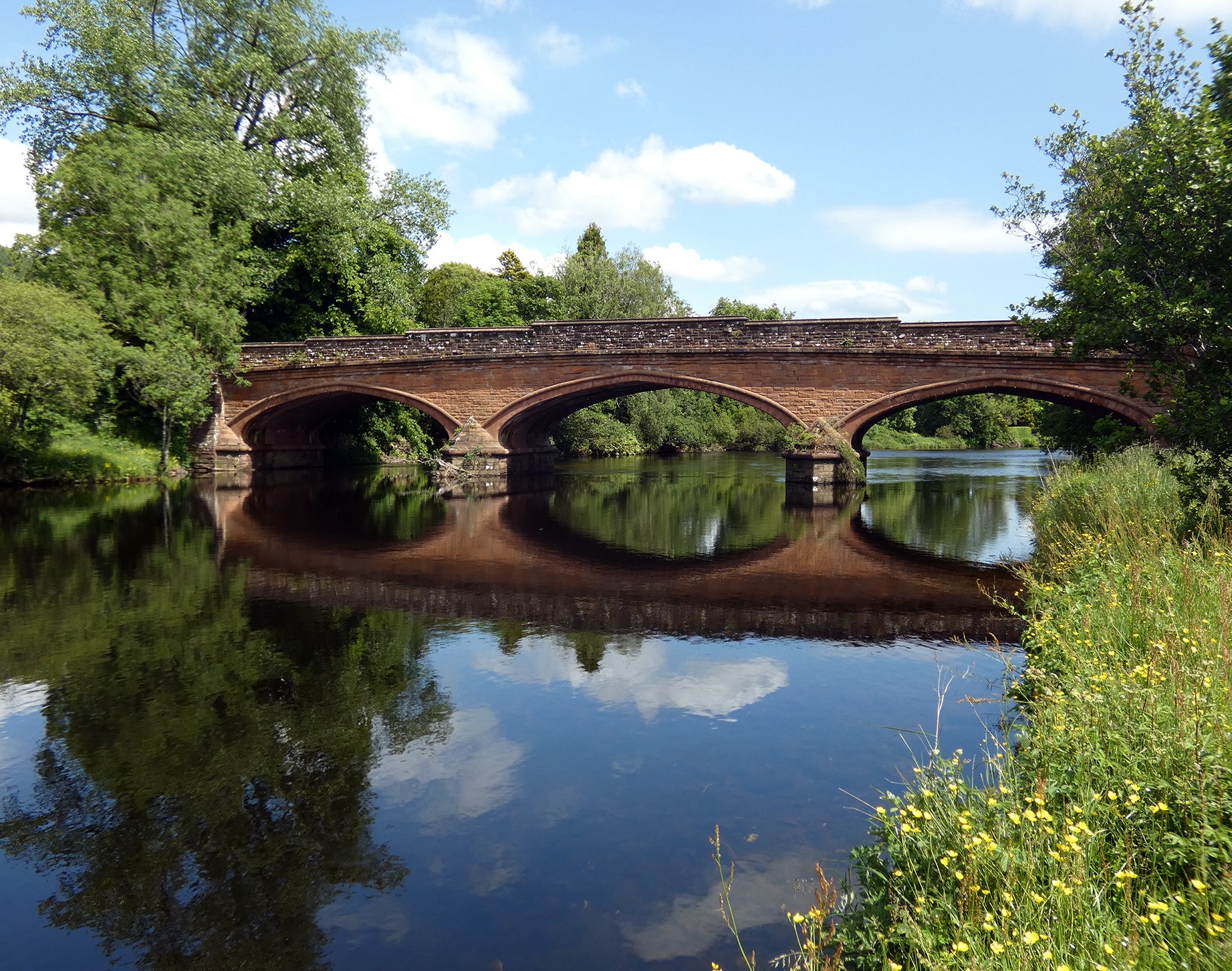 Stone bridge over a calm river with trees and blue sky in the background