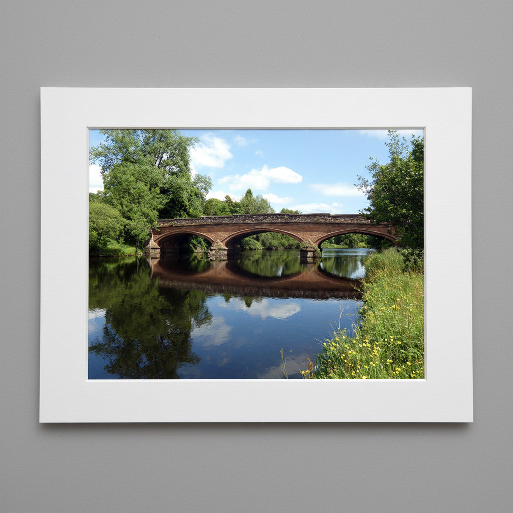 Framed photograph of a bridge over a calm river with trees on either side.